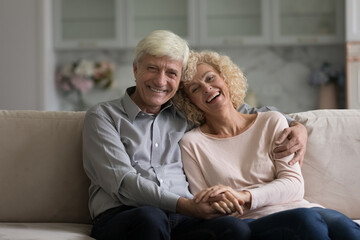 Portrait of optimistic married elderly couple in love staring for cam seated on sofa at modern home. Smiling older spouses looking posing for camera. Carefree well-being retired life, happy marriage