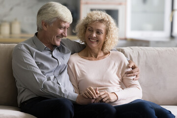 Attractive older couple in love talking sit on couch at modern home. Optimistic elderly wife and grey-haired husband relaxing in cozy living room enjoy conversation, spend leisure together feel happy