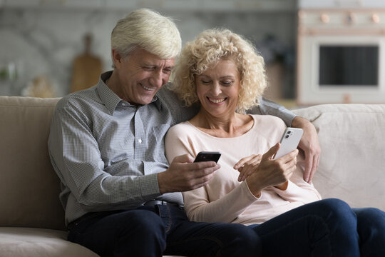 Positive mature 70s spouses spend free time with two smartphones, sit on sofa staring at cellphone screens learn new devices, gadgets feel happy. Older generation using modern tech, application usage