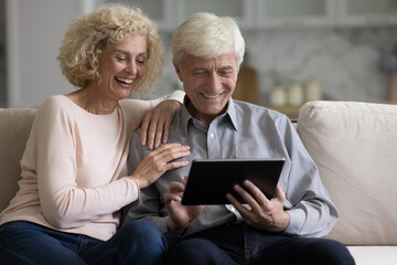 Retired couple spends free time use digital tablet. Senior grey-haired husband his aged blond wife enjoy internet usage, buying goods ordering services through e-commerce retail websites. Technology
