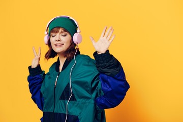 a happy, joyful woman in a green jacket and hat enjoys music standing in pink headphones on a yellow background raising her hand up
