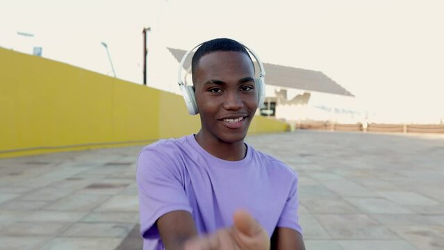 Front view of smiling young happy african american dancer man in headphones dancing outdoors. Millennial student guy having fun listening to music alone against yellow wall. Youth culture concept
