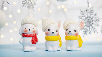A Christmas card, three ceramic rabbits in knitted hats and a Santa Claus hat stand on a blue background, among Christmas balls and snowflakes, with garland lights. the concept of Christmas.
