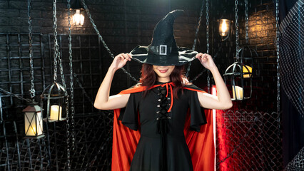 Naklejka premium Happy gothic young woman in witch halloween costume with hat standing and smiling in halloween theme.