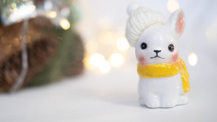 A Christmas card, a small ceramic rabbit in a knitted hat and scarf stands against the background of a blurred Christmas tree with garland lights.