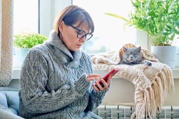 Woman in sweater sitting in armchair near window with heating radiator and sleeping cat