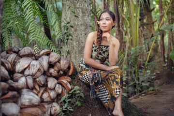 A Balinese girl in batik clothes sits among the remains of coconuts.
