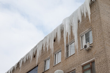 Big icicles on the roof of a townhouse on a snowy winter day among thaw. Cleaning the roofing from snow and icicles.