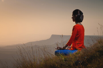 young caucasian woman meditates in the lotus position in the grass in the mountains near the cliff in the setting sun