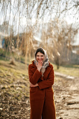 Young attractive girl in orange coat, headscarf and scarf, laughs at camera, covering her mouth with one hand, other covers her stomach, autumn season.