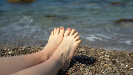 Female feet on the background of sea waves. A woman lies on the shore with small stones close to the water. Holiday concept by the sea.