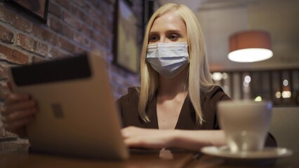A business woman in a medical mask is talking on a video link using a tablet in a cafe. Working phone call, remote work. Coronavirus epidemic.