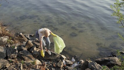 A woman removes rubbish among the stones near the lake. The woman takes the scattered rubbish and puts it in a trash bag.
