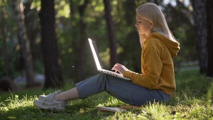 Naklejka na ściany i meble A young blonde woman in casual clothes with a laptop sits on the green grass. The sun illuminates the laptop. Books are lying nearby.