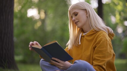 Young pretty woman reads an adventure novel with interest in nature. Woman leafs through a book on green grass.
