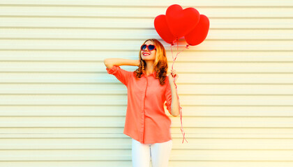 Portrait of happy smiling young woman with bunch of red heart shaped balloons wearing shirt, sunglasses on white background