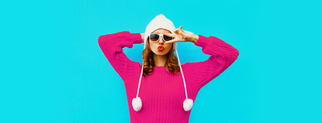 Portrait of stylish young woman blowing her red lips wearing pink sweater and hat with pom pom on blue background