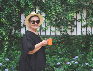  healthy senior asian woman wearing black blouse, sunglasses and hat, holding orange color cup of coffee in the garden with purple flowers, smiling and looking at camera.