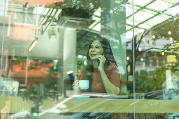 Obraz na płótnie Canvas A girl takes a phone call while having a cup of coffee with light reflection at the window. Lady sitting alone having a conversation through mobile phone beside a shop window with light reflection.