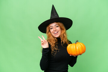 Young caucasian woman costume as witch holding a pumpkin isolated on green screen chroma key background smiling and showing victory sign