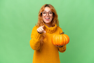 Young caucasian woman holding a pumpkin isolated on green screen chroma key background surprised and pointing front