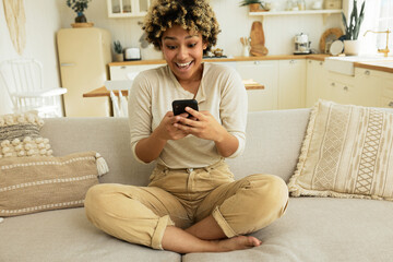 Obraz premium Cute afro woman with surprised happy excited face looking at smartphone screen sitting on couch in living-room against kitchen furniture, astonished with unbelievable news, special offer, sales ad