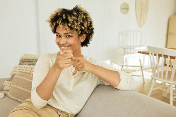 Smiling tricky cunning sly female of 20s with afro hairstyle and dark skin sitting on couch in...