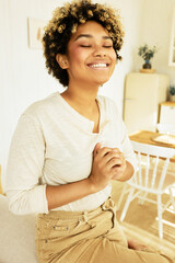Vertical portrait of happy cheerful young african american girl sitting on back of couch with folded hands, making wish with closed eyes and joyful smile, dressed in casual beige clothes