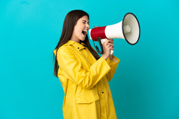 Teenager Brazilian girl wearing a rainproof coat over isolated blue background shouting through a megaphone