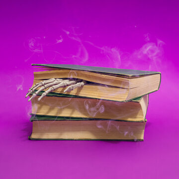 Halloween or horror scene with old books,  skeleton hand and smoke on purple background. Horror story concept.