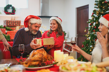 Happy family celebrating Christmas dinner at home. Granddaughter with gift box given to grandfather wearing Santa hats.Merry Christmas or New Year's eve concept