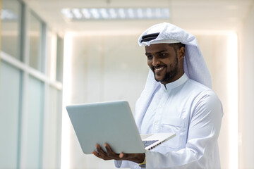 Muslim Businessman in Traditional White Kandura Standing in His Modern Office and Using Laptop Computer