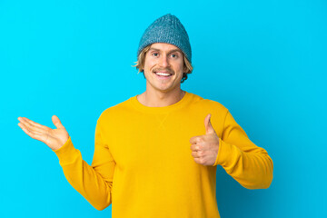 Naklejka premium Handsome blonde man isolated on blue background holding copyspace imaginary on the palm to insert an ad and with thumbs up
