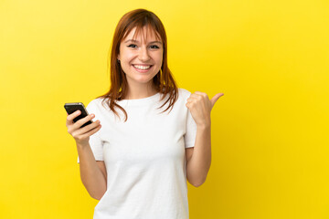 Fototapeta premium Redhead girl isolated on yellow background using mobile phone and pointing to the lateral