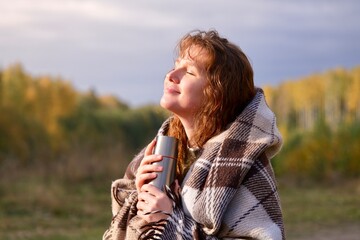 Portrait of beautiful girl, young happy woman is drinking tea or coffee, hot beverage from thermo cup, thermos bottle, covering herself in plaid, cozy blanket in natural golden autumn park or forest - obrazy, fototapety, plakaty