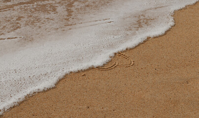 Close up on the foam of a wave occulting a love heart hand drawn in the sand of a beach.