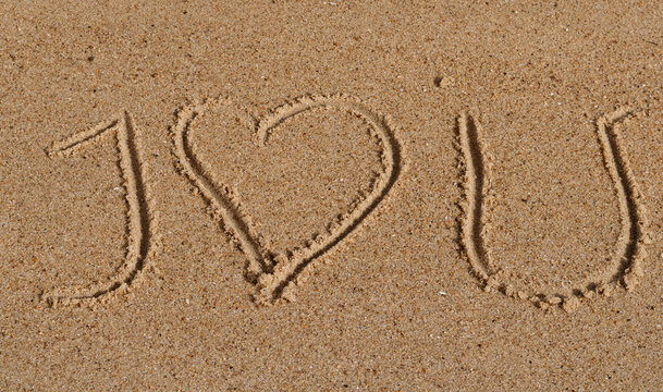 Close up on a message hand drawn in the sand of  a beach saying " I love you". The word love is replace by a heart.