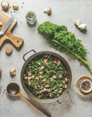 Poster Cooking pot with stewed kale with mushrooms on kitchen table with spoon and ingredients, top view © VICUSCHKA
