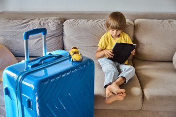 A sad child is sitting on a sofa using a tablet and waiting for departure. Family relocation,...