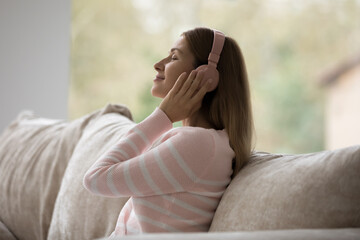 Internet-based music streaming services user, modern wireless tech usage. Young calm woman in headphones listens affirmations, audiobook or favourite song seated on sofa resting at home looks peaceful