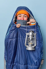 Vertical shot of pleased Asian woman wrapped in warm sleeping bag during frosty weather holds...