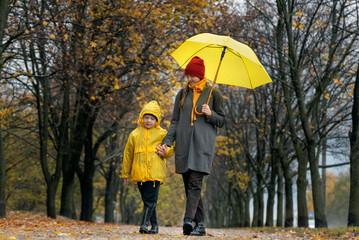 Mom and her child are walking in the park on rainy autumn day. Mom and son with big yellow umbrella.