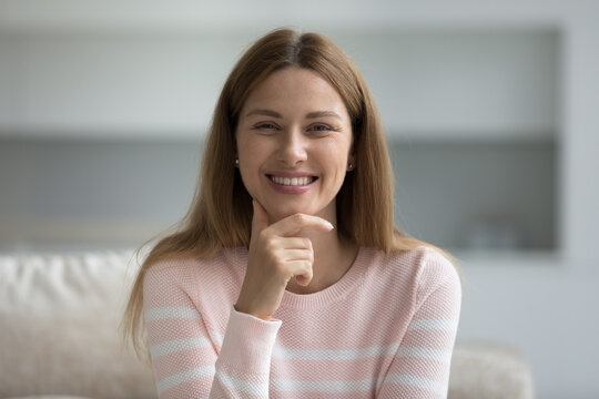 Head shot beautiful woman sit on sofa looking at camera relaxing on sofa at home, happy homeowner portrait, millennial female with wide candid smile advertises dental clinic services. Natural beauty