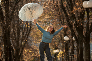 Laughing young woman holds transparent umbrella on outstretched arms. Cheerful girl in blue sweater on autumn park