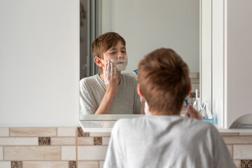Little brunette boy applies shaving foam to face. Reflection in the mirror. Daily morning procedures