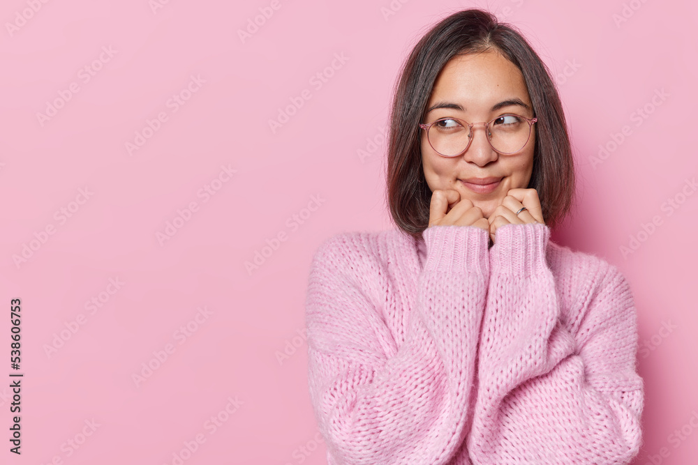 Wall mural Horizontal shot of pretty Asian woman keeps hands under chin looks tenderly aside being deep in thoughts wears round spectacles knitted winter jumper isolated over pink background blank space