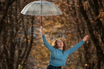 Student laughs and holds transparent umbrella on outstretched arms.Cheerful girl in blue sweater, autumn park background