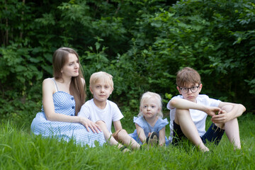 Naklejka na ściany i meble Young mother with three children sits on green lawn. Beautiful woman with small daughter and two sons resting on grass