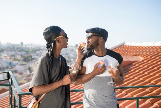 Portrait of happy African gay couple eating cakes. Two men in sunglasses standing together with glasses of champagne and one man feeding another man cake. Concept of happiness in love of LGBT couples