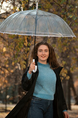 Smiling young woman with long hair whirling under transparent umbrella in autumn park. Woman smiles directly into camera